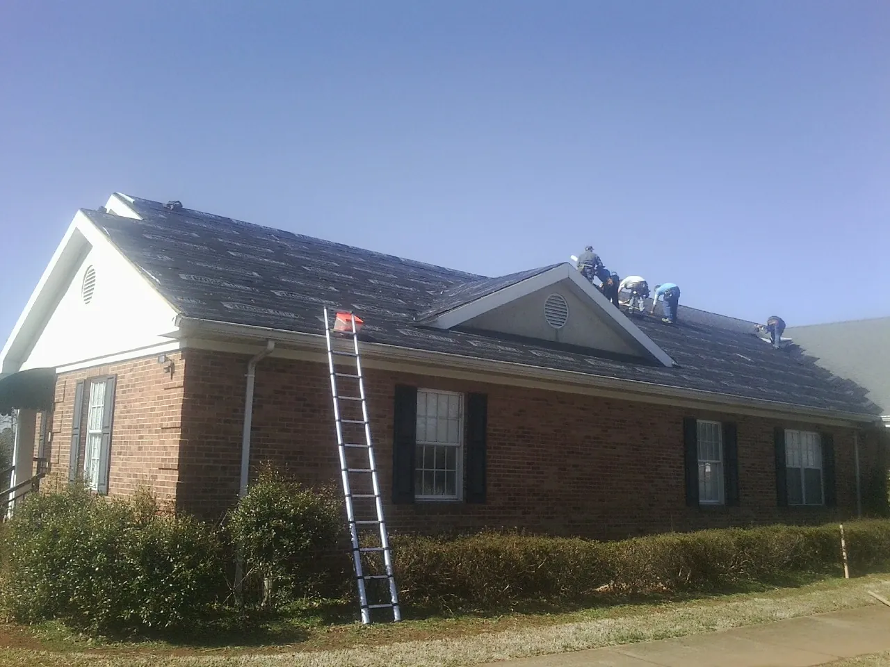 Crew tearing shingles off a brick ranch roof with a ladder set — Emergency Roof Repair work by our crew in New Hill