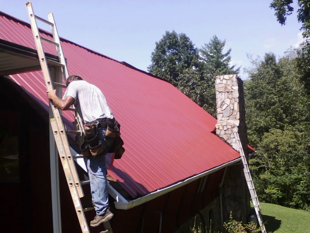 Roofer on a ladder beside a red metal roof and stone chimney — Soffit & Fascia Repair by Roofing Brothers in New Hill